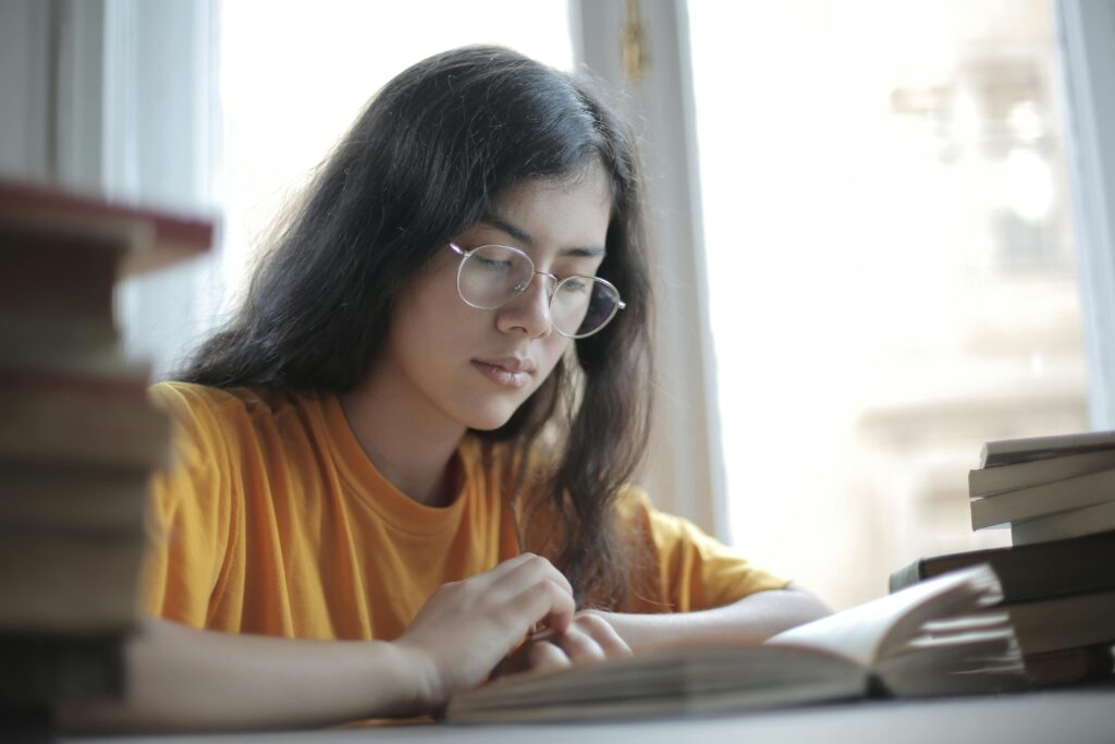 Low angle of diligent ethnic female student wearing casual t shirt and eyeglasses sitting at table with stacks of books and preparing for exam